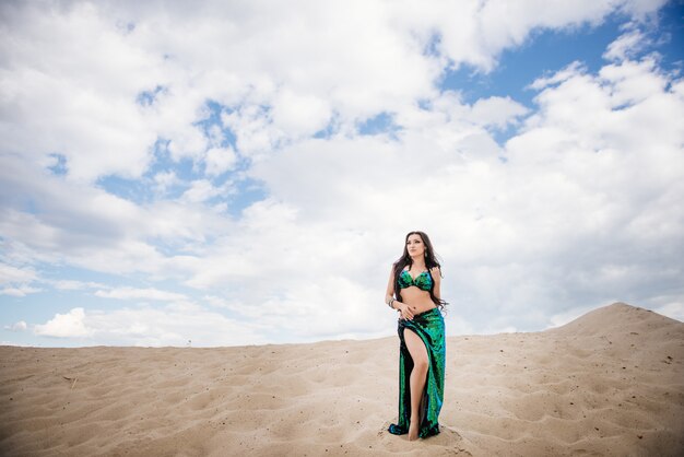 Dançarina do ventre em um terno brilhante preto e verde no deserto no contexto da areia.