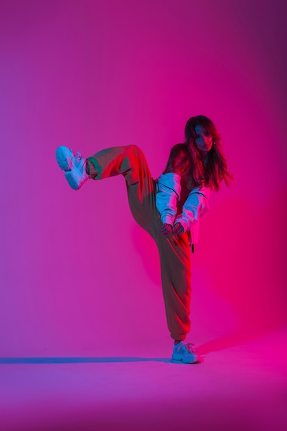 Dançarina elegante jovem com roupas da juventude da moda dos esportes em tênis da moda fica em uma perna no estúdio com uma luz rosa brilhante. Menina moderna dançando na sala com a cor de néon multicolorida.