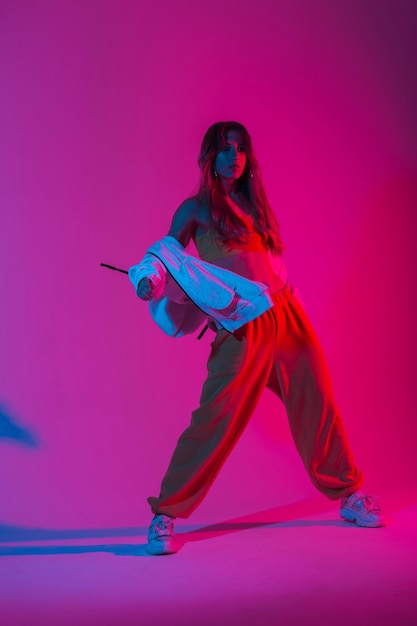 Dançarina muito jovem em um agasalho de treino de moda jovem em tênis elegantes, dançando hip-hop no estúdio com luz de néon rosa brilhante. Garota legal gosta de dançar com cores multicoloridas no estúdio.