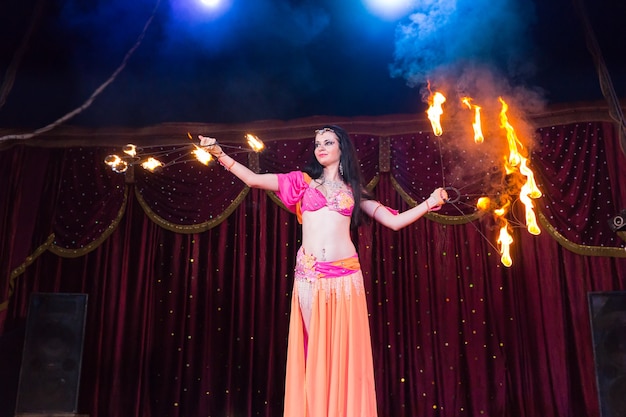 Dançarino do fogo exótico de cabelos escuros girando em um aparelho de bastão flamejante no palco iluminado por holofotes