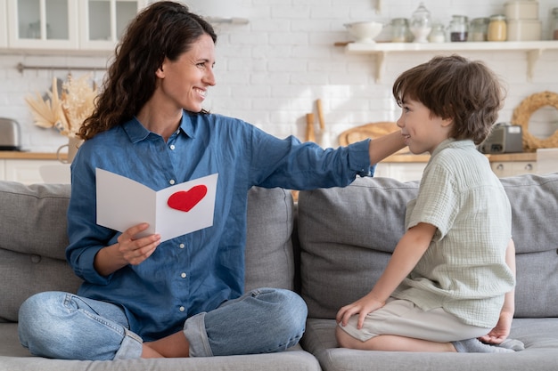 Dankbare Mutter berührt süßes Kind Sohn Haare halten Postkarte mit Grüßen am Muttertag sitzen auf der Couch zu Hause