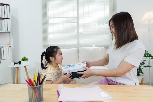 Dankbares asiatisches Mädchen mit blauer Geschenkbox, das der Mutter ein Muttertagsgeschenk macht. Fröhliche Mutter und süße Tochter feiern Geburtstag und umarmen sich zu Hause auf dem Sofa. Fröhliches Familienkonzept