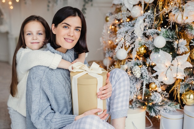 Dankbares kleines weibliches Kind umarmt ihre Mutter, die Geschenk gab, wundervolle unvergessliche Zeit zusammen verbringt, Weihnachten feiert. Brunettefrau und -tochter suchen nach anwesenden Geschenken unter Tannenbaum