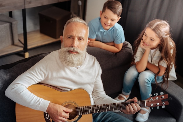 Dankbares Publikum. Inspirierter älterer Mann, der Gitarre spielt und für seine Enkel singt, während sie ihm mit Bewunderung zuhören