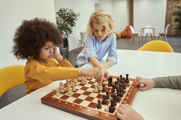Dar consejos a los niños pequeños inteligentes que discuten el movimiento mientras están sentados a la mesa y juegan al ajedrez en