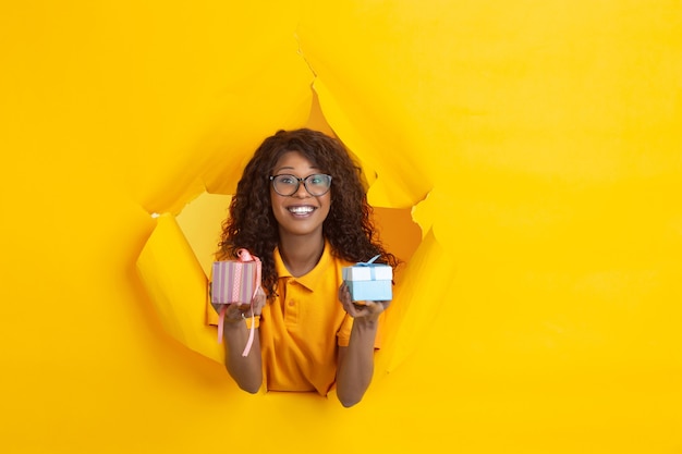 Dar presentes. Jovem afro-americana alegre em fundo de papel amarelo rasgado, emocional, expressivo. Rompendo, descoberta. Conceito de emoções humanas, expressão facial, vendas, anúncio.