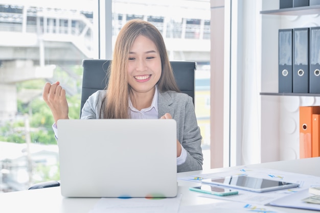 Das asiatische Geschäft, das mit Laptop arbeitet und den Erfolg hat, der Arme anhebt, erhalten nach gute Nachrichten