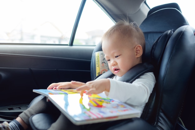 Das asiatische Kind, das im haltenen Autositz sitzt und genießen, Buch zu lesen