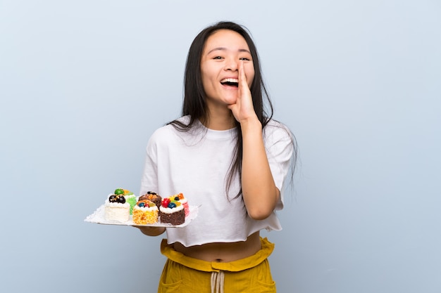 Das asiatische Mädchen des Jugendlichen, das viele verschiedene Minikuchen schreiend mit dem breiten Mund hält, öffnen sich