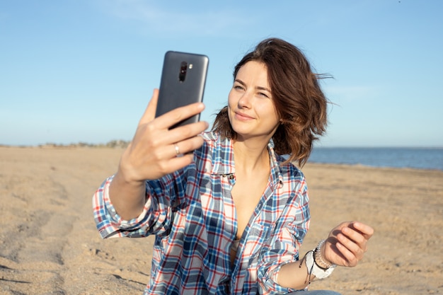 Das atmosphärische Lebensstilfoto der jungen schönen dunkelhaarigen Frau im Hemd und in den Jeans im Freien macht Selfie am Strand