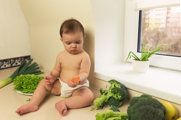 Das Baby in der Windel sitzt auf dem Küchentisch. Ein Kind spielt und hat Spaß mit frischem Bio-Gemüse und Obst.