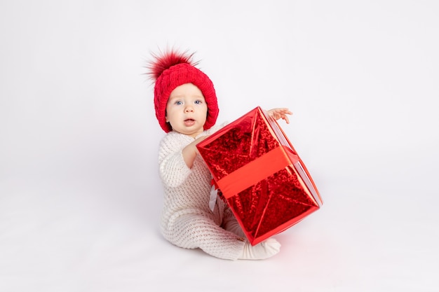 Das Baby mit dem roten Hut und dem Geschenk auf weißem lokalisiertem Hintergrund, Raum für Text, Konzept des neuen Jahres und Weihnachten