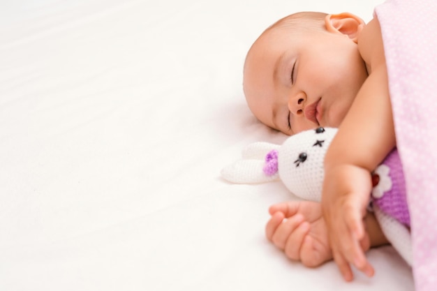 Das Baby schläft auf dem Bett mit seinem Lieblingsspielzeug unter der Decke. Ein Baby im Traum umarmt einen gestrickten Hasen und konzentriert sich selektiv auf das Gesicht des Kindes