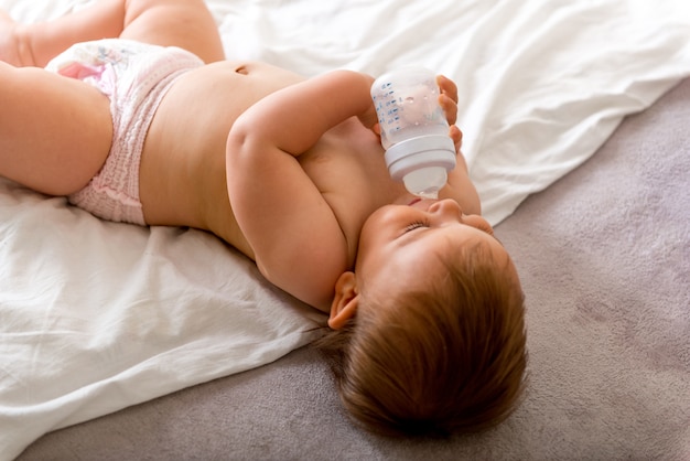 Das Babykleinkind, das auf das weiße Bett legt, lächelt und trinkt Wasser von der Plastikflasche