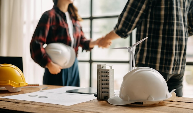 Das Bauteam schüttelt Hände und begrüßt den Start eines neuen Projektplans hinter einem gelben Helm auf dem Schreibtisch im Bürozentrum, um sich über ihr Bauprojekt zu beraten