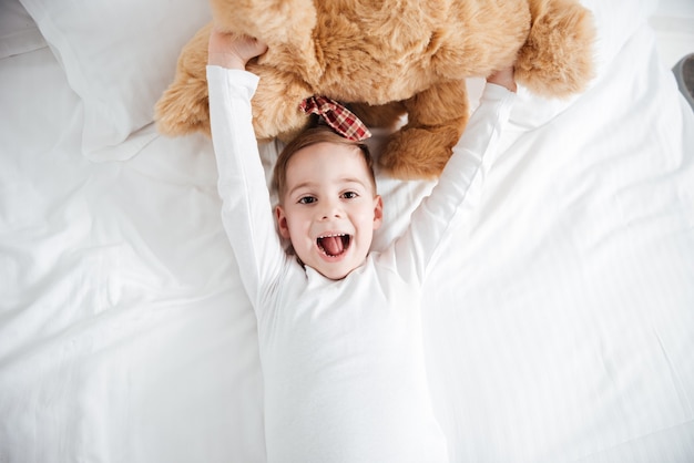 Das Bild des fröhlichen kleinen Jungen liegt zu Hause auf dem Bett und hält Teddybären. Schau nach vorne.