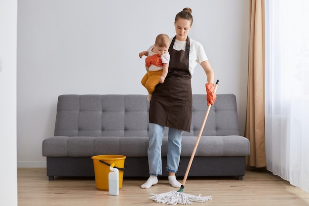 Das Bild einer traurigen, verärgerten, müden Frau, die eine braune Schürze mit Wischmopp und eine kleine Kleinkindtochter in den Händen trägt, die im Wohnzimmer putzt und traurig nach unten schaut, braucht Ruhe