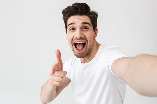 Das Bild eines aufgeregten, glücklichen jungen Mannes, der isoliert über der weißen Wand posiert, macht ein Selfie, indem die Kamera auf Sie zeigt.