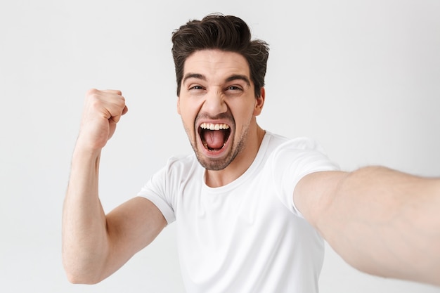 Das Bild eines aufgeregten, glücklichen jungen Mannes, der isoliert über der weißen Wand posiert, macht ein Selfie mit der Kamera und macht Siegergeste.