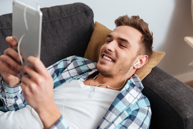 Das Bild eines fröhlichen Borstenmannes, der in einem Hemd in einem Käfigdruck gekleidet ist, liegt auf dem Sofa zu Hause und verwendet einen Tablet-Computer