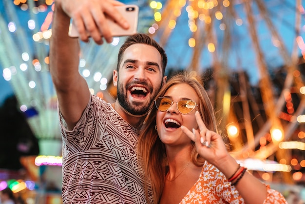 Das Bild eines glücklichen, süßen, fröhlichen, liebevollen Paares, das im Vergnügungspark im Freien spazieren geht, nimmt ein Selfie-Handy, das Frieden zeigt.