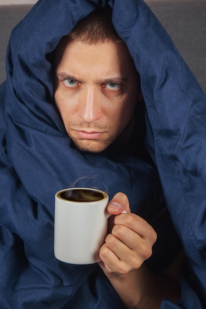 Das Bild eines jungen Mannes sitzt auf einem Bett und ist mit einer dunkelblauen Decke bedeckt. Er hält eine weiße Tasse Tee. Der Typ schaut in die Kamera. Er ist ernstes Coronavirus und kaltes Konzept