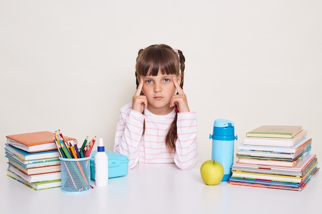 Das Bild eines traurigen, ernsten kleinen Schulmädchens mit Zöpfen, das ein gestreiftes Hemd trägt, hält die Finger an den Schläfen und blickt in die Kamera, die am Schreibtisch sitzt, umgeben von Büchern und anderem Schulmaterial