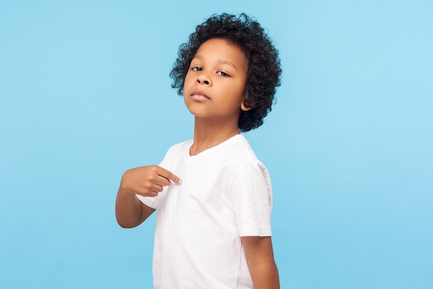 Das bin ich Porträt eines lockigen Vorschuljungen in T-Shirt, der stolz und arrogant auf sich selbst zeigt, mit ernsthaftem, hochmütigem Ausdruck, egoistisches Kind, Studioaufnahme isoliert auf blauem Hintergrund