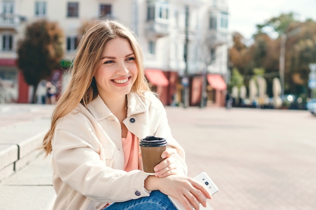 Das blonde Mädchen, das auf den Schritten mit einem Tasse Kaffee sitzt, schaut weg und lächelt, Kopienraum