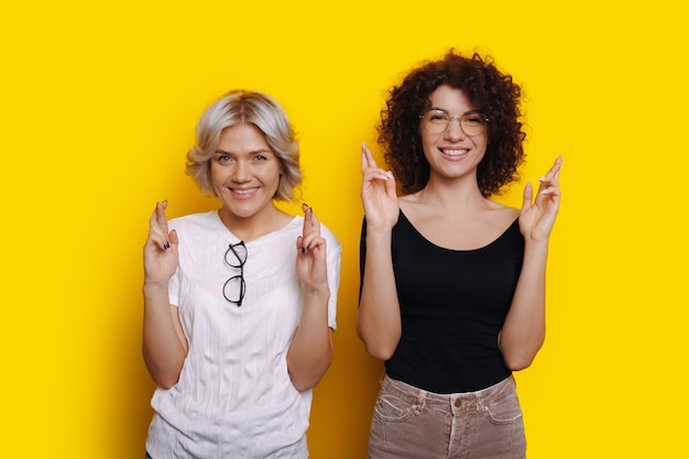 Das blonde Mädchen und ihre lockige Freundin lächeln liebevoll und kreuzen ihre Finger an einer gelben Wand