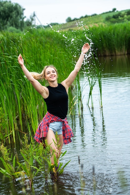 Das blonde Mädchen vergnügt sich im Fluss in Gummistiefeln auf dem Land