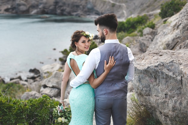 Das Brautpaar über die Natur in den Bergen am Wasser. Anzug- und Kleidfarbe Tiffany. Gehen Sie Hand in Hand.