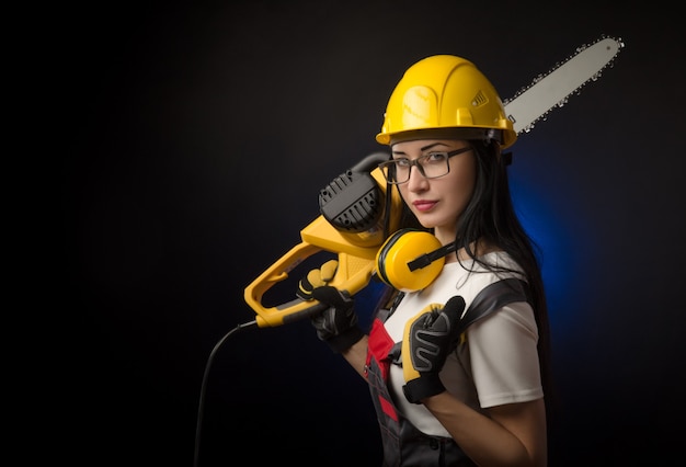 Das brünette Mädchen in Spezialkleidung und ein Arbeiter in einem Helm posiert auf schwarzem Hintergrund mit einem Arbeitswerkzeug (Motorsäge)