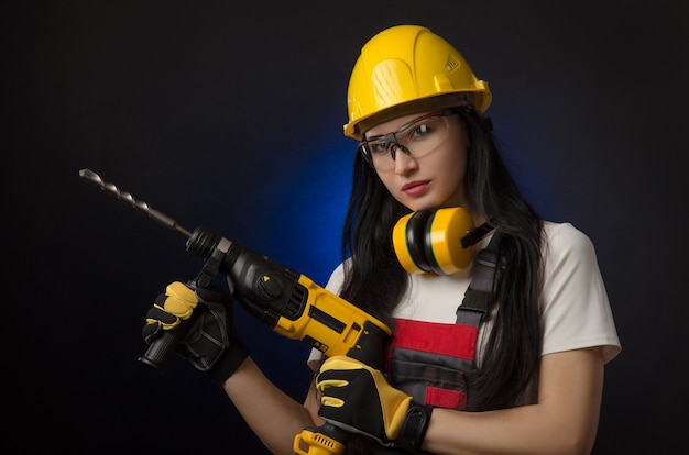 Das brünette Mädchen in Spezialkleidung und ein Arbeiter in einem Helm posiert auf schwarzem Hintergrund mit einem Arbeitswerkzeug (Perforator, Bohrer, Schlaghammer)