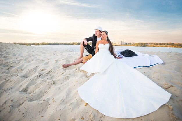 Das charmante Mädchen des Hochzeitspaares in einem weißen Kleid und im positiven Mann im Hut sitzen auf dem weißen Sand am Strand auf Hintergrund