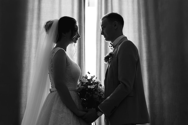 Das erste Treffen von Braut und Bräutigam Weißes Hochzeitskleid, grauer Anzug und stilvoller Blumenstrauß Jungvermählten halten sich an den Händen und sehen sich an