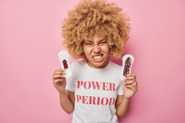 Das Foto einer gereizten jungen Frau hat kritische Tage und hält zwei Servietten, die mit Blut schmutzig sind, die Zähne zusammenbeißt und die Menstruation hasst, die in einem lässigen T-Shirt mit weißer Aufschrift einzeln auf rosafarbenem Hintergrund gekleidet ist