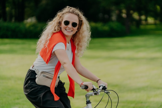 Das Foto einer lockigen Europäerin hält die Hände am Lenker von Fahrradreisen im Freien im Park trägt ein lässiges T-Shirt mit Schattierungen und eine schwarze Hose lächelt positiv und drückt das Glück aus Erholung der Menschen
