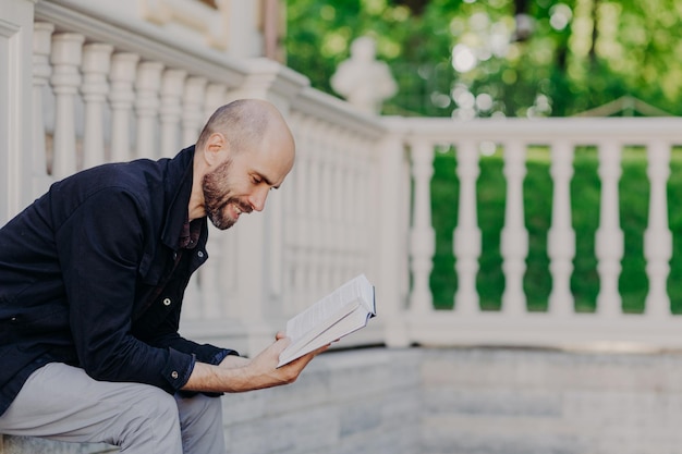 Das Foto eines kahlen bärtigen Mannes mittleren Alters lehnt sich eng an ein Buch an und hat einen positiven Ausdruck. Er sitzt auf einem weißen Balkon und hat dunkle Stoppeln