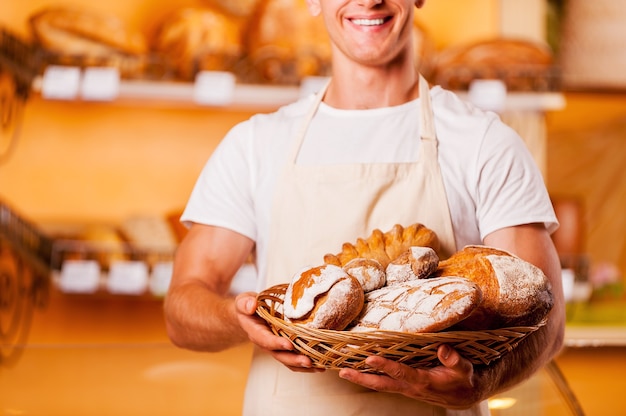 Das frischeste Brot für Sie. Abgeschnittenes Bild eines jungen Mannes in Schürze, der einen Korb mit Backwaren hält und lächelt, während er in der Bäckerei steht