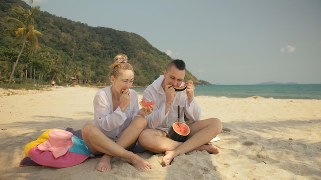 Das fröhliche Liebespaar, das Wassermelonenscheiben auf tropischem Sandstrand hält und isst