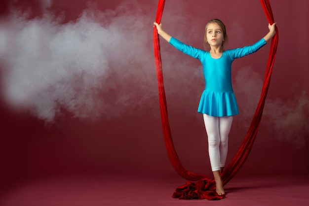 Das furchtlose hübsche kleine Mädchen in einem blauen Turnanzug zeigt ein rotes Stunt-Luftband, umgeben von Rauch auf einem blass-blassen Hintergrund. Konzept der fortgeschrittenen Akrobatenturner