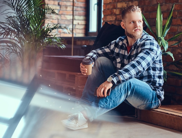 Das Ganzkörperbild eines blonden Hipster-Männchens trinkt Kaffee in einem Raum mit Loft-Interieur.