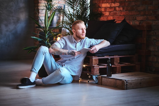 Das Ganzkörperbild eines blonden, stylischen Mannes in Fleecehemd und Jeans mit einem Tablet-PC in einem Wohnzimmer.