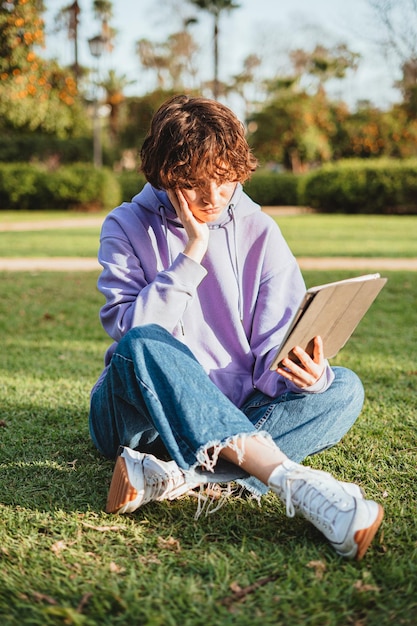 Das gelangweilte Mädchen, das einen lila Hoodie trägt, benutzt ein digitales Tablet, während es auf dem Gras sitzt.