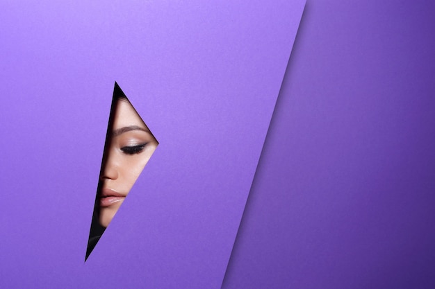 Das Gesicht einer jungen schönen Frau mit einem hellen Make-up schaut durch ein Loch in violettem Papier.