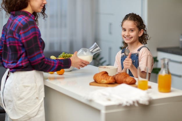 Das glückliche kleine Mädchen zeigt die Daumen beim gesunden Frühstück zusammen mit der Mutter in der Küche.