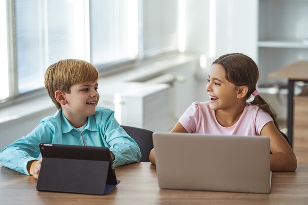 Das glückliche Mädchen und ein Junge mit Gadgets sitzen am Tisch