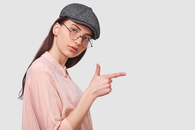 Das horizontale Porträt einer aufrichtigen jungen Frau mit rosafarbenem Hemd und runder transparenter Brille zeigt mit dem Zeigefinger etwas an einem leeren Kopierraum für Werbung, die ernsthaft aussieht