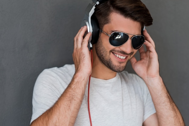 Das ist mein Lieblingslied! Hübscher junger Mann mit Kopfhörern, der die Musik hört und lächelt, während er vor grauem Hintergrund steht